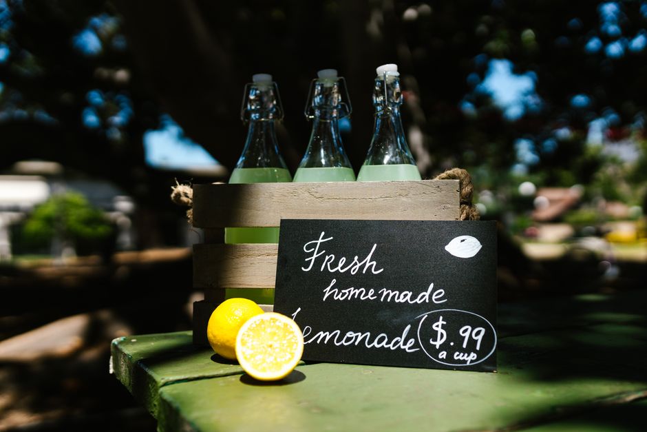 Caixa de garrafas de vidro com limonada em uma mesa ao ar livre, com preço de R$0,99 por copo.